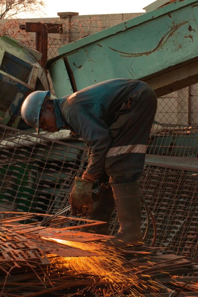 A blue-collar worker welding metal in a scrap yard, creating sparks.